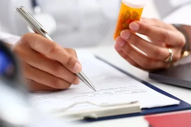 Physician holding prescription medicine and writing patient notes in a chart.